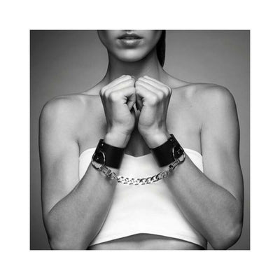 Black wide vegan leather cuffs connected by a silver chain worn on a woman's wrists with a white tube top.