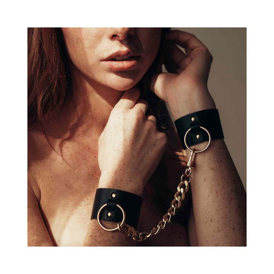 Close-up of a woman wearing black wide vegan leather cuffs connected by a gold chain on her wrists.