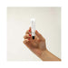 Hand with red nail polish holding a small white tube of Bijoux Indiscrets Slow Sex Oral Sex Balm against a plain background.