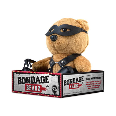 Plush teddy bear wearing black faux leather harness, boots, and eye mask, holding a small flogger in Bondage Bearz branded box.