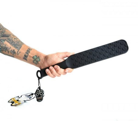 Hand holding a black silicone paddle with raised skull designs and a hanging loop with a skull keychain attached.