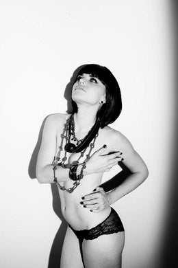 Black and white photo of a woman in lace underwear and layered necklaces posing against a plain wall.