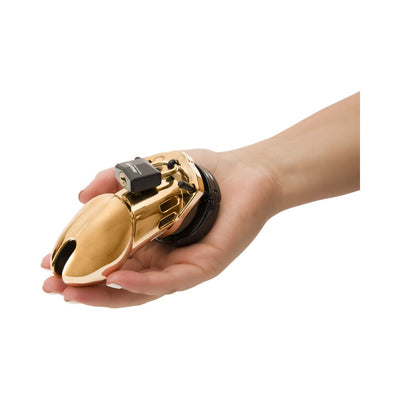 Hand holding a gold-colored male chastity device with a black lock and base ring on a white background.