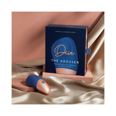 Deia The Arouser silicone external stimulator in blue and rose gold with packaging on silky fabric background.