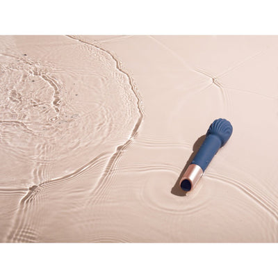 Blue silicone wand vibrator with rose gold base resting in shallow water with ripples on a light surface.