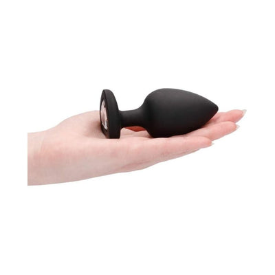 Black extra large silicone butt plug with a diamond-shaped gem at the base held in a hand on a white background.