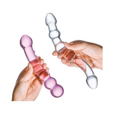 Hands holding two double-ended glass dildos, one clear and one pink, with smooth and textured designs.
