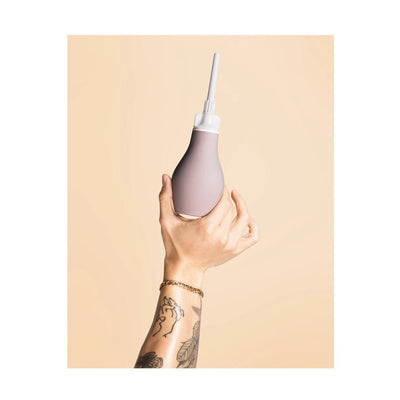 Hand holding a soft silicone anal douche with a removable white nozzle against a beige background.