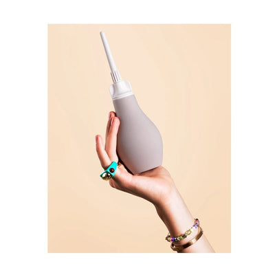 Hand holding a gray silicone anal douche with a white nozzle against a beige background.