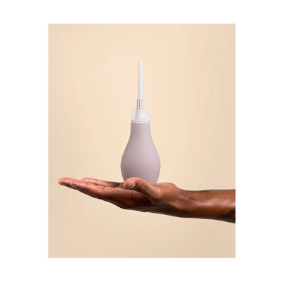 Hand holding a soft silicone anal douche with a white removable tip against a beige background.