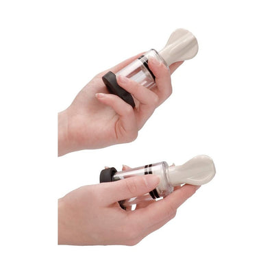 Hands holding two white and transparent electro nipple twisters with black accents on a white background.