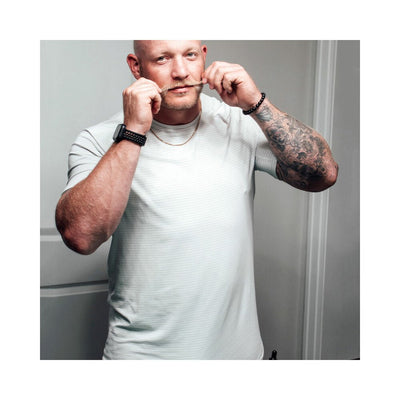 Man with a trimmed beard and tattoos styling his mustache, wearing a white shirt and black wrist accessories.