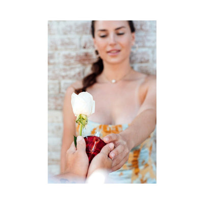 Woman receiving a white rose and a red diamond-shaped perfume bottle in an outdoor setting
