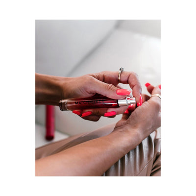 Close-up of hands with bright coral nails holding and spraying Eye of Love Matchmaker Pheromone Perfume in a sleek red bottle.
