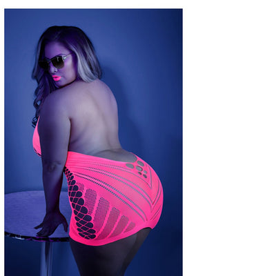 Plus size model wearing neon pink glow net halter dress with open back and geometric patterns, posing by a small round table.