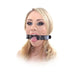 Blonde woman wearing a black beginner open mouth gag with adjustable strap and tongue out against white background.
