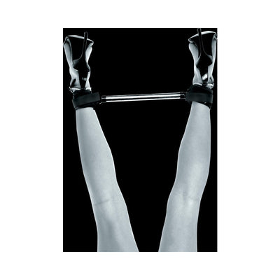 Black metal spreader bar with adjustable ankle cuffs worn with high-heeled shoes on raised legs against black background.