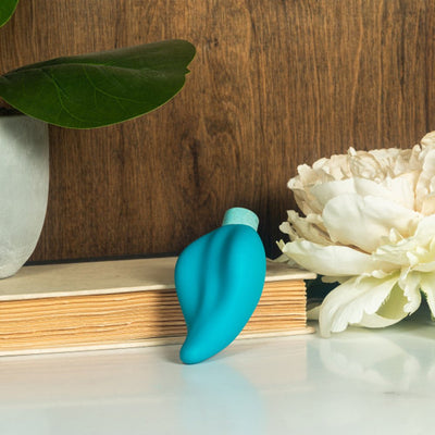 Blush Gaia Eco Caress in aqua with smooth, curved design displayed on a book next to a plant and white flower.