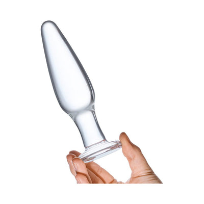 Hand holding a clear glass anal plug with a tapered shape and flared base against a white background.