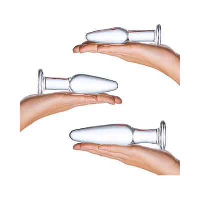 Three clear glass anal training plugs of varying sizes held in hands against a white background.