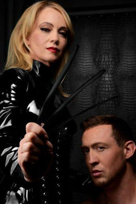 Woman in black latex holding three black silicone urethral sounds with a man kneeling beside her against a dark background.