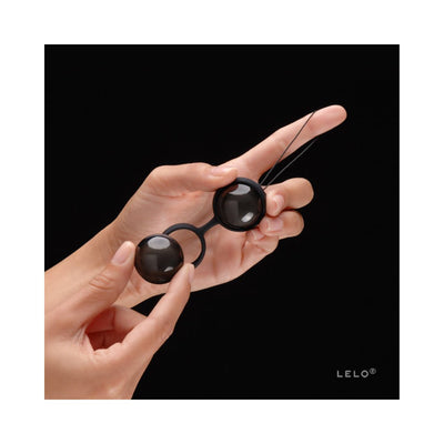 Hands holding glossy black LELO Beads Noir connected by a silicone harness against a black background.