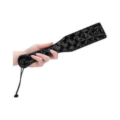 Hand holding a black luxury paddle with a textured diamond pattern and a wrist strap on a white background.
