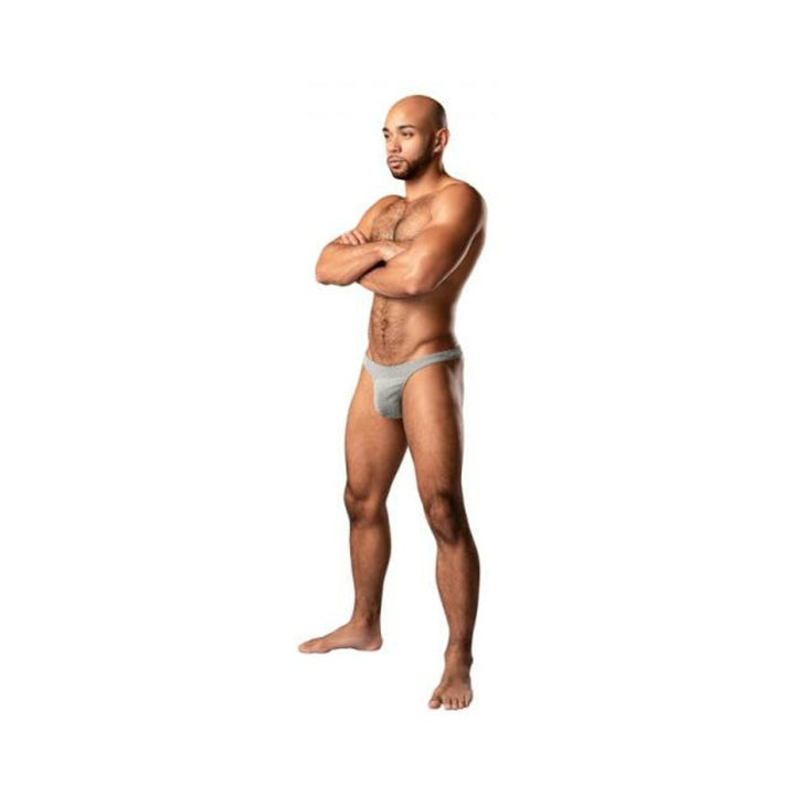 Male model wearing grey weave Male Power Labyrinth Bong Thong with low cut waist and t-back design, standing with arms crossed.