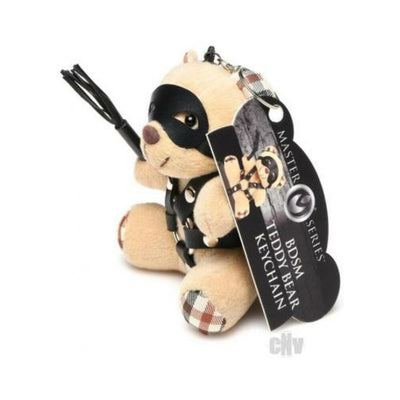 Small plush teddy bear keychain with light peach fur, black faux leather harness, eye mask, and mini flogger accessory.