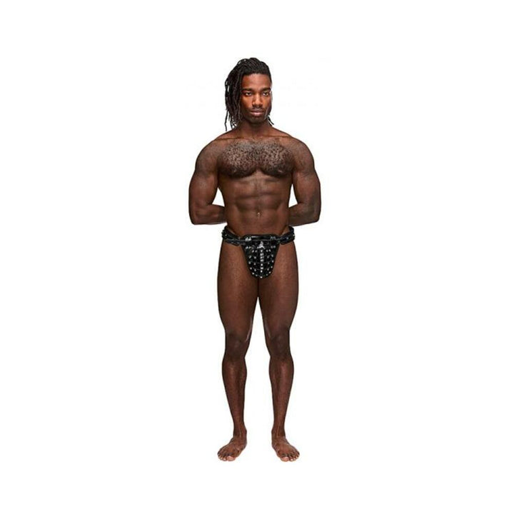 Athletic man wearing black leather thong with studded cage pouch and chastity lock, standing against white background.
