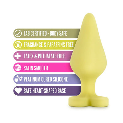 Yellow silicone anal plug with heart-shaped base shown alongside colorful icons highlighting body-safe, fragrance-free, latex-free, satin smooth, and platinum-cured features.