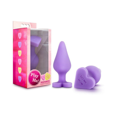 Purple silicone anal plug with heart-shaped base engraved "Do Me Now" next to pink packaging featuring candy heart designs.