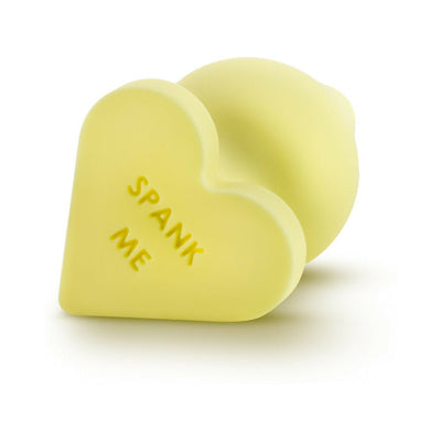Yellow heart-shaped silicone anal plug with "SPANK ME" text on the base, smooth satin finish, flexible design.