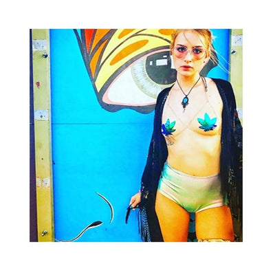 Woman wearing green to black flip sequin weed leaf pasties with high-waisted shorts and a black cover-up against colorful mural background.