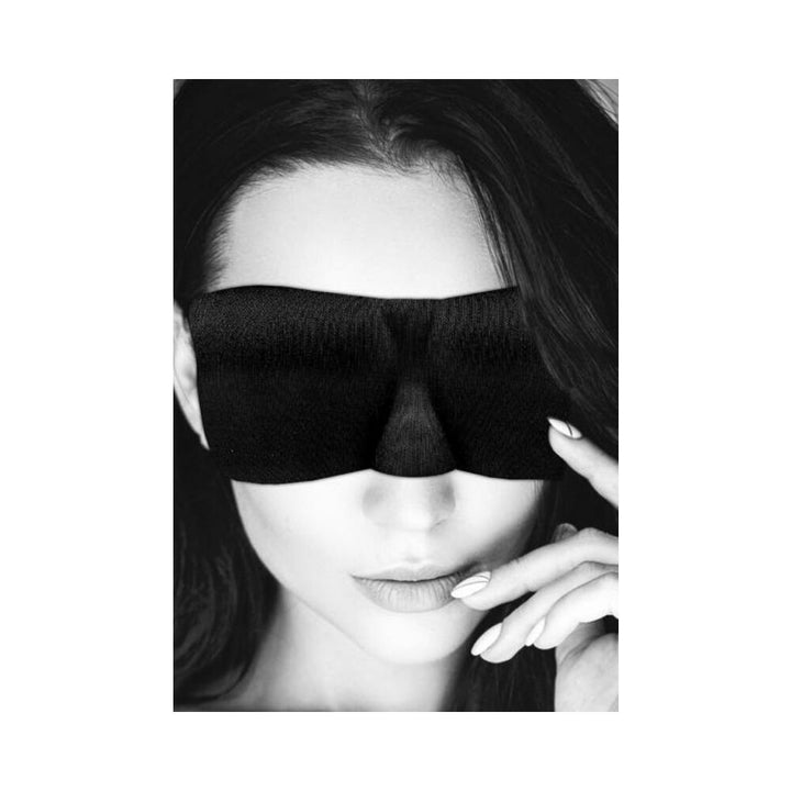 Woman wearing a black satin curvy eye mask with elastic straps, touching her face gently.