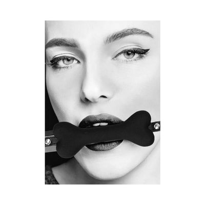 Close-up black and white image of a woman wearing a black silicone bone-shaped gag with adjustable leather straps.