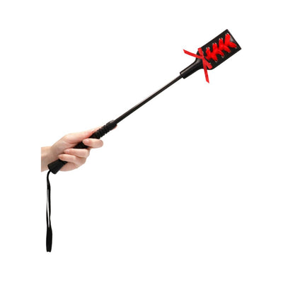 Hand holding a black crop with a rectangular tip decorated with red lace ribbon detailing.