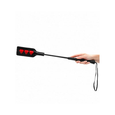 Hand holding a black crop with three red heart shapes on the paddle and a wrist strap.