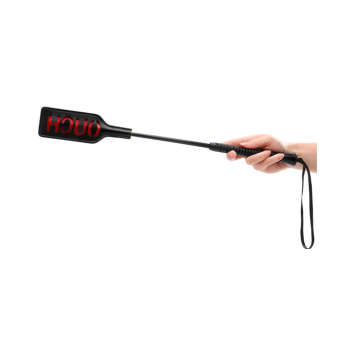 Hand holding a black Ouch! Crop with a short handle and rectangular tip embossed with the word OUCH in red.