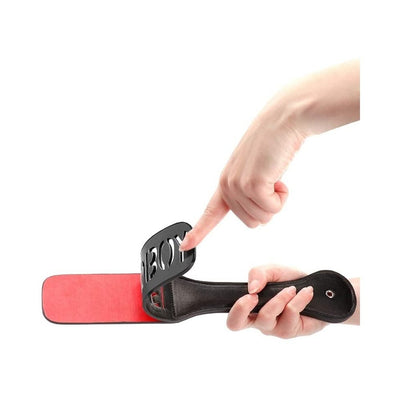 Black leather paddle with red striking surface and cut-out "BAD BOY" text held by hands on white background.