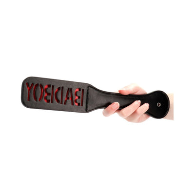 Hand holding black leather paddle with red backing and cut-out "BAD BOY" lettering on paddle face.