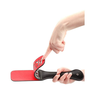 Hand holding a black leather paddle with red striking surface and cut-out reversed lettering spelling "BITCH" on the paddle face.