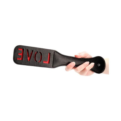 Hand holding black leather paddle with cut-out reversed LOVE letters revealing red backing underneath.