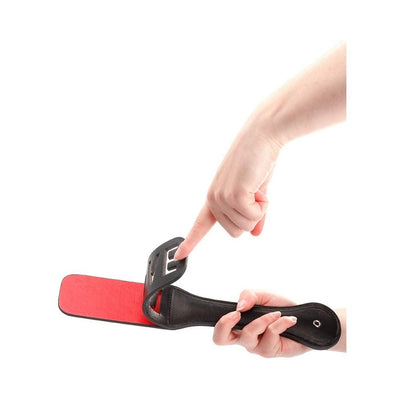 Black leather paddle with red striking surface and cut-out reversed "LOVE" design held by hands on white background.