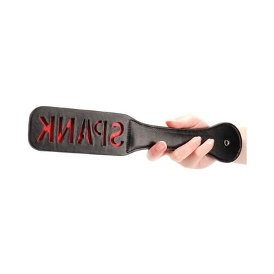 Hand holding black leather paddle with red "SPANK" cutout lettering on paddle face against white background.