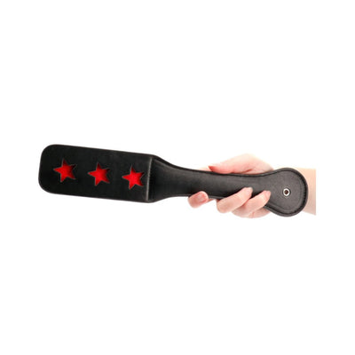 Hand holding black leather paddle with three star-shaped cutouts revealing red backing beneath.