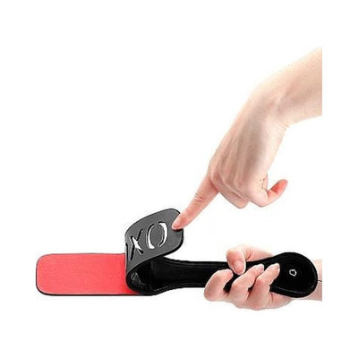 Black leather paddle with red striking surface and cut-out XOXO design held and pointed at by a hand.