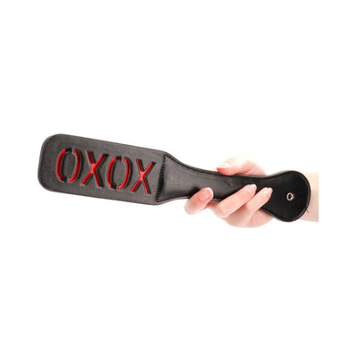 Hand holding black leather paddle with red "XOXO" cutout design and stitched edges against white background.