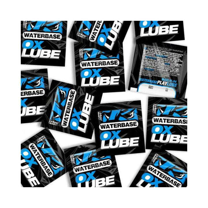 Multiple black and blue single-use packets of Oxballs Waterbase Oxlube lubricant scattered on a white background.