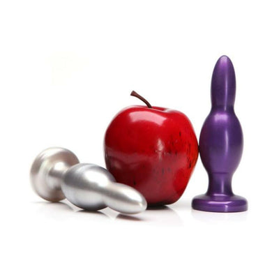 Silver and purple metal training plugs displayed next to a red apple for size comparison on a white background.